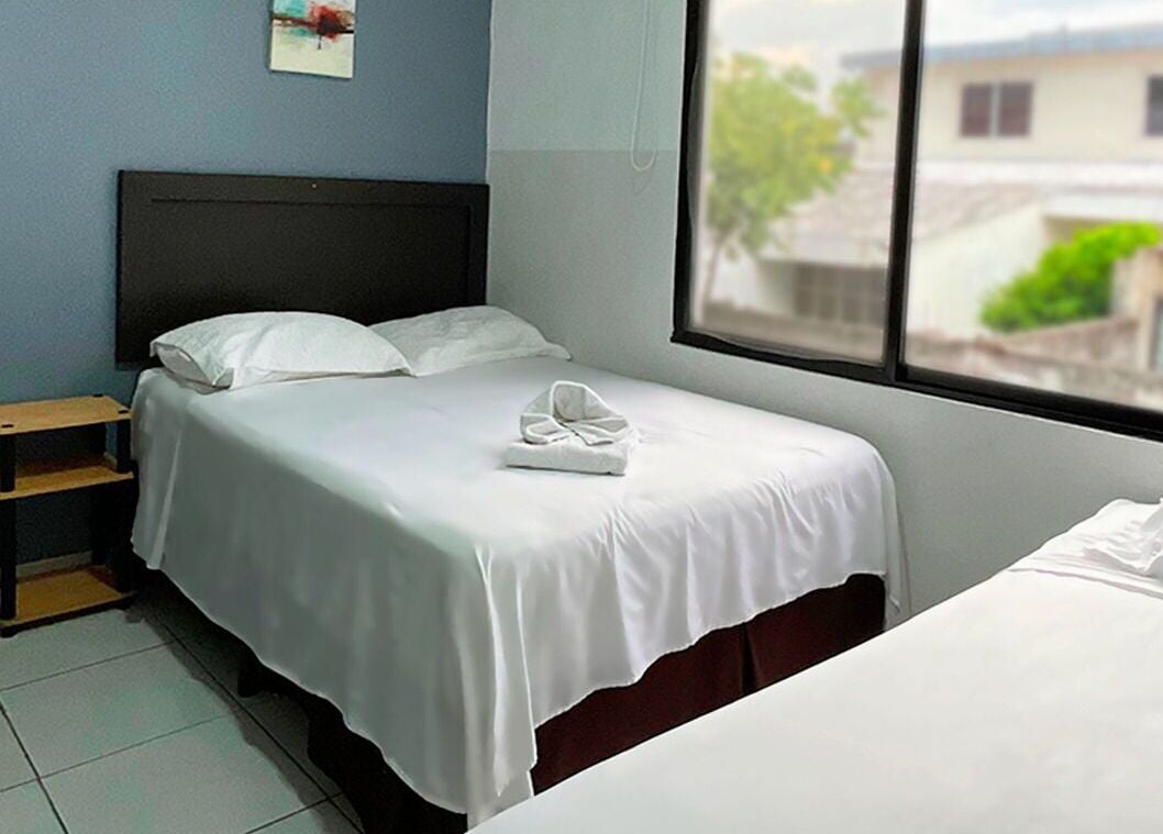 Une chambre d’hôtel avec un lit simple, une table de chevet, une fenêtre donnant sur des immeubles et une lampe fixée au mur.