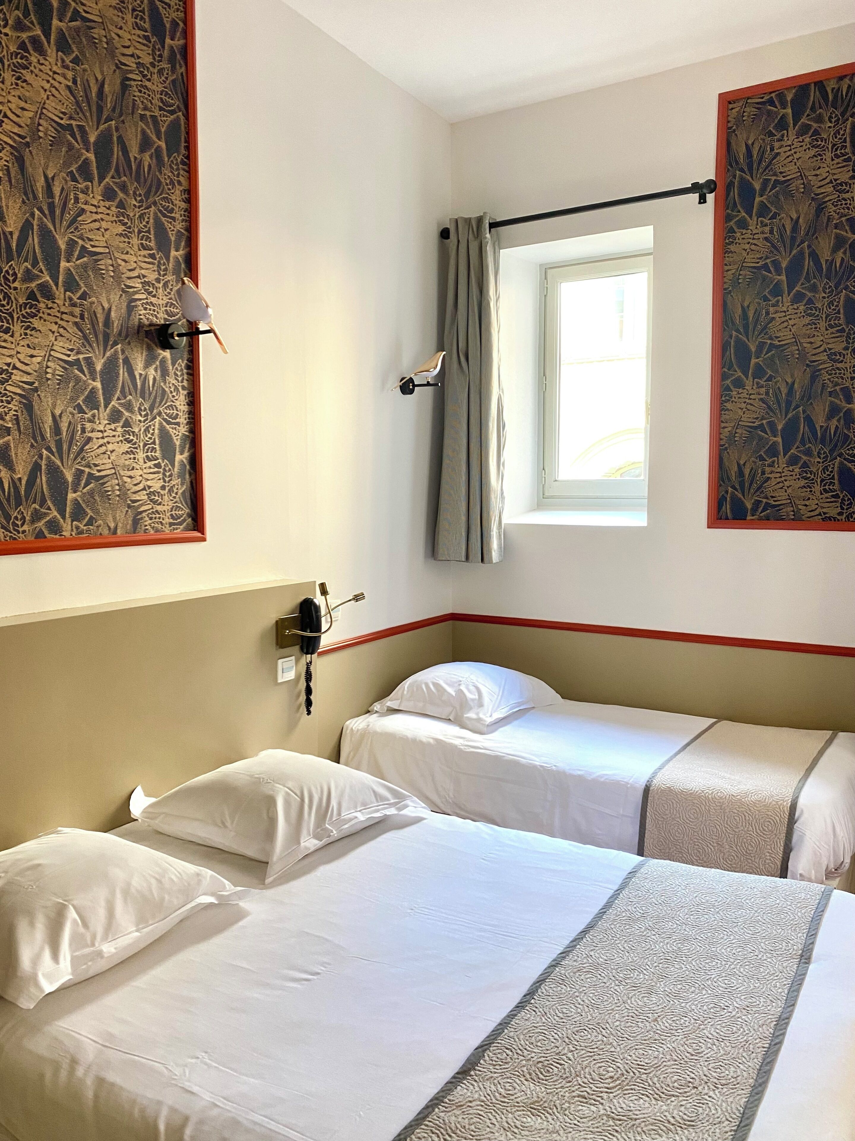 Ett hotellrum med två sängar, ett fönster med gardiner, en telefon som sitter fast på väggen och dekorativa paneler.