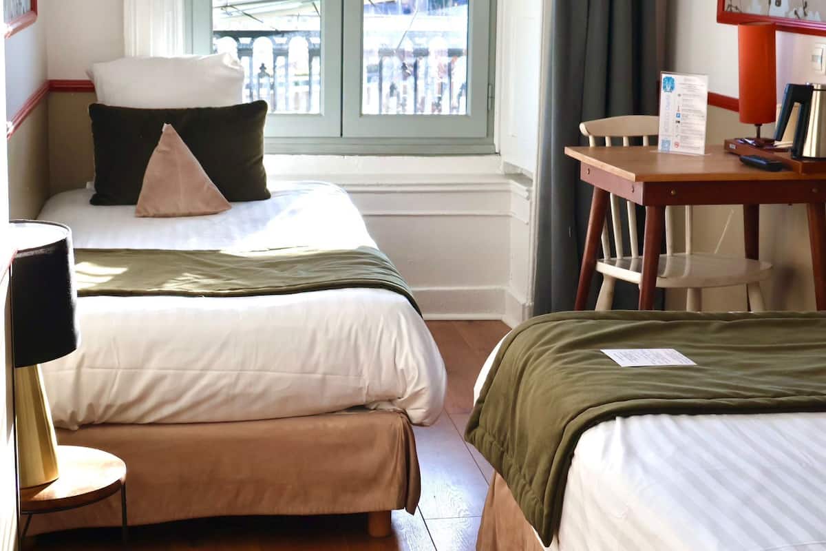 A hotel room with two beds, a small table with a kettle, a chair, and a window with a view of the outside.