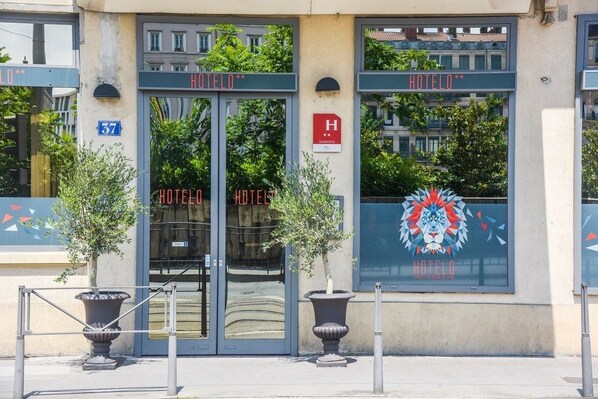 L’entrée d’un hôtel, avec un double jeu de portes, une enseigne indiquant « HÔTEL » et un logo représentant la tête d’un lion.