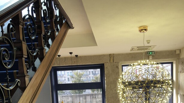 A hotel lobby with a staircase, a large window with a view of the street, a modern chandelier, and a seating area with a plant and a yellow chair. - HOTELO Lyon Charité (Lyon)