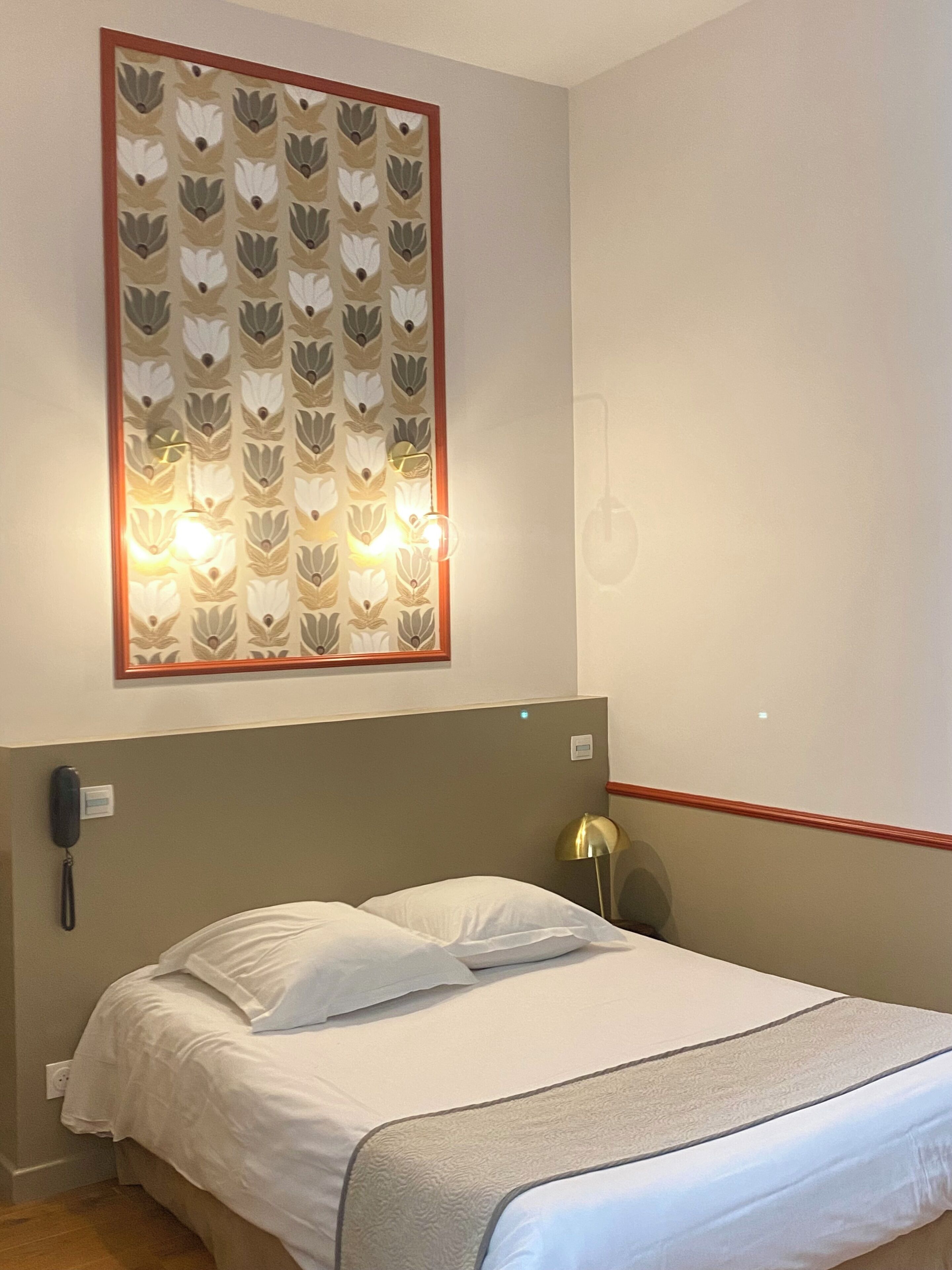 Une chambre d’hôtel équipée d’un lit, d’un téléphone fixé au mur et d’un luminaire orné d’un motif floral.