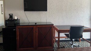 A hotel room with a flat-screen TV mounted on the wall, a wooden desk, a black office chair, and a microwave on a cabinet. - American Inn (Columbia)