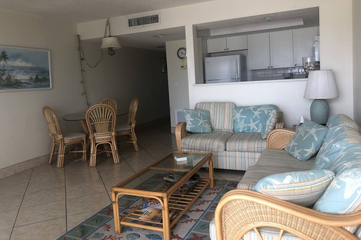 A living room with a sofa, chairs, a glass coffee table, and a kitchen area in the background.