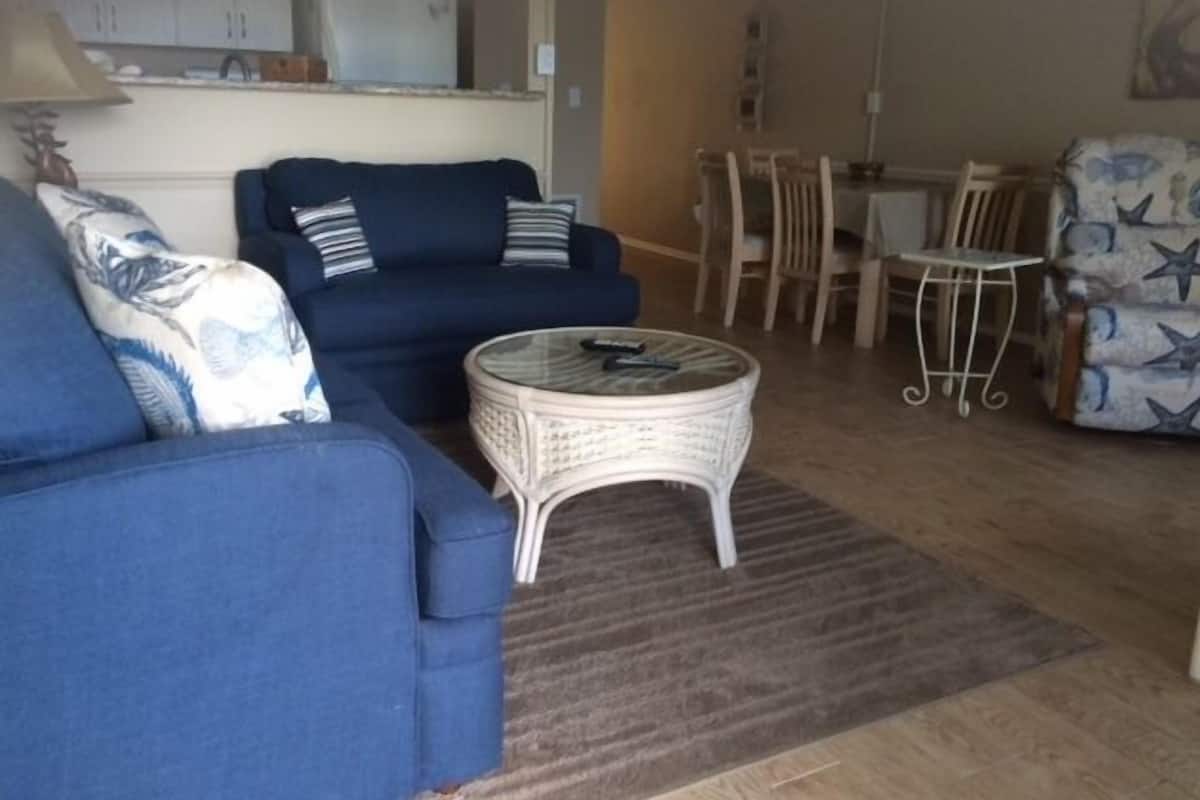 A living room with a blue sofa, a round wicker coffee table, and a dining area with a small table and chairs.