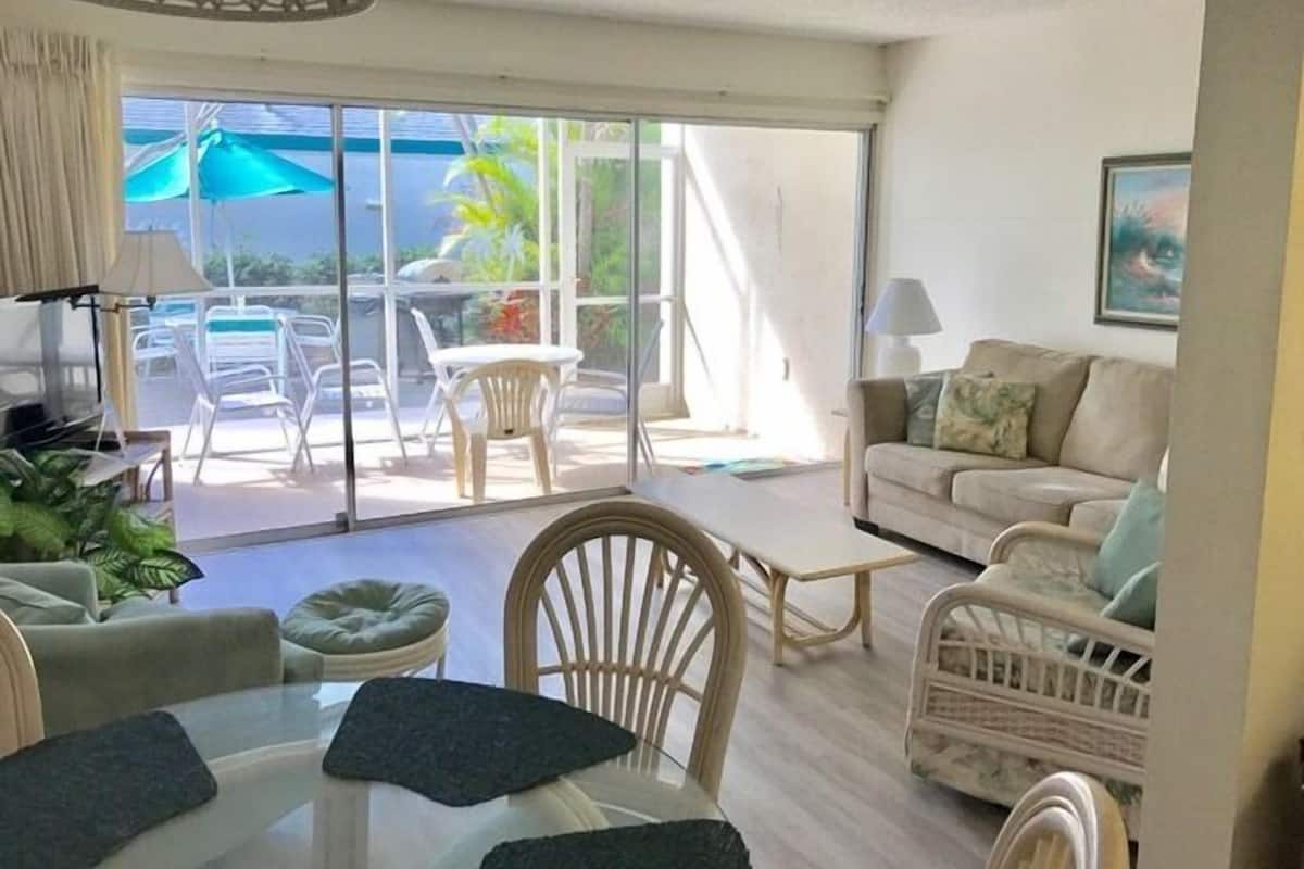 A spacious living area with a large sliding glass door leading to an outdoor patio. Inside, there is a round table with chairs, a sofa, and a wicker chair.