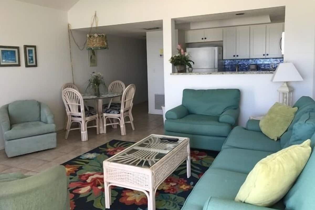 A living room with a sofa, chairs, and a coffee table. There is a dining area with a table and chairs. A kitchen is visible in the background.
