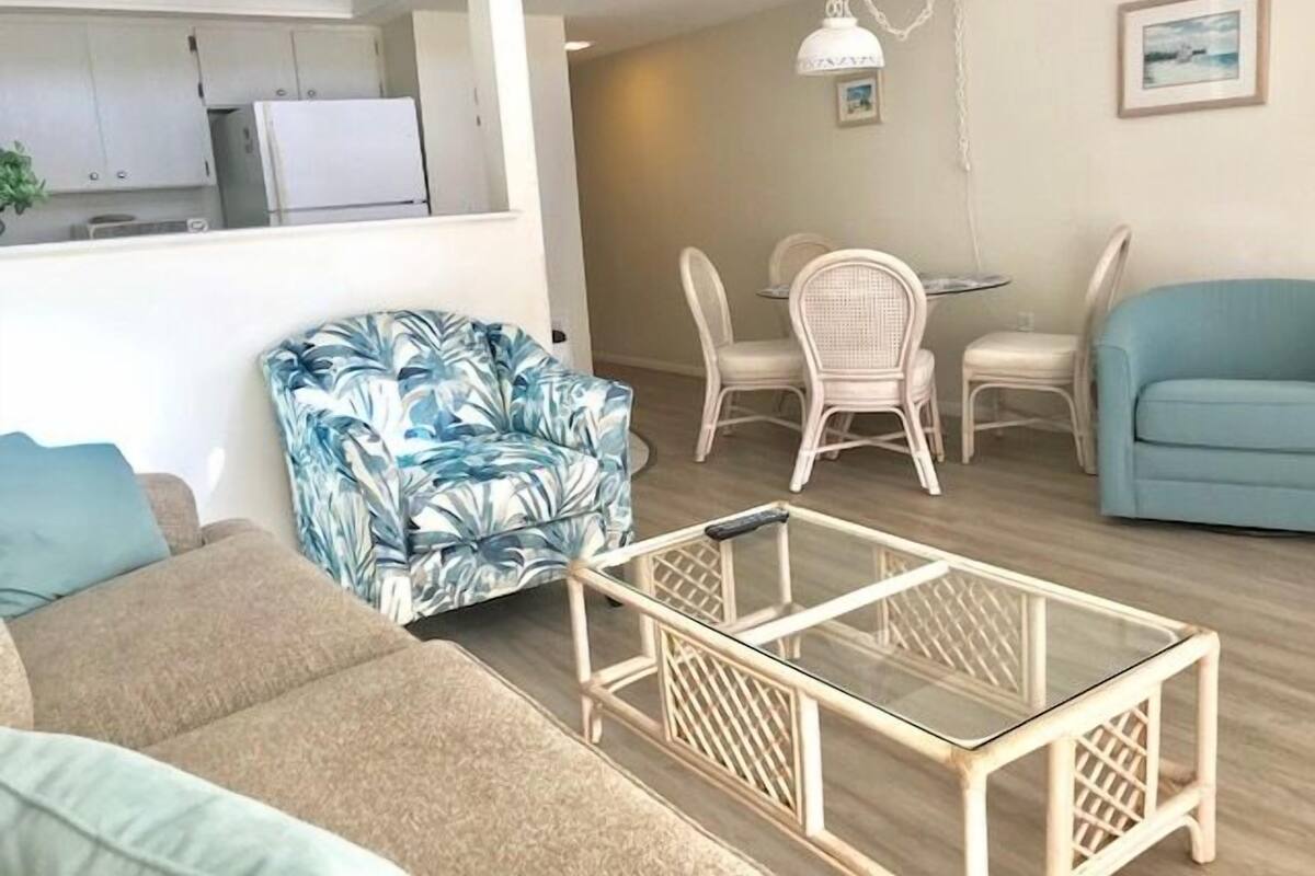 A living room with a sofa, a patterned armchair, a glass coffee table, and a dining area with a table and chairs.
