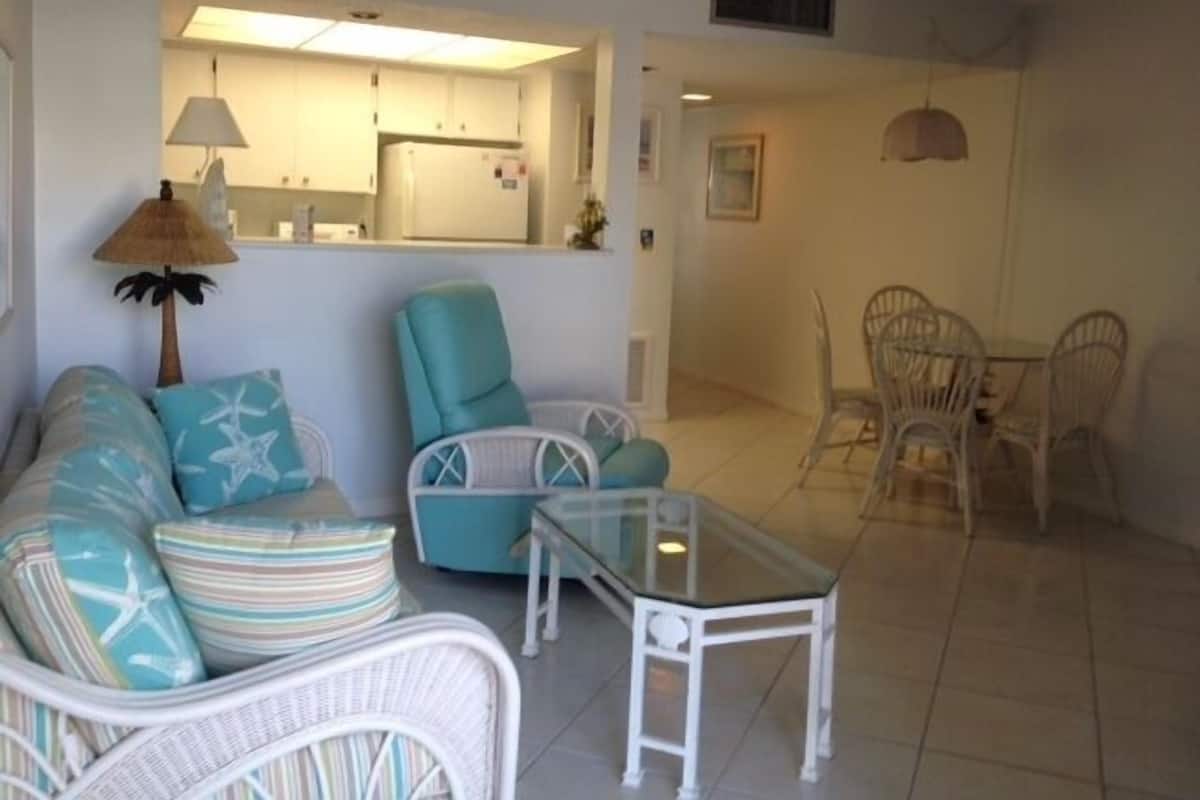 A living room with a sofa, armchair, and glass coffee table. There is a dining area with chairs and a table.
