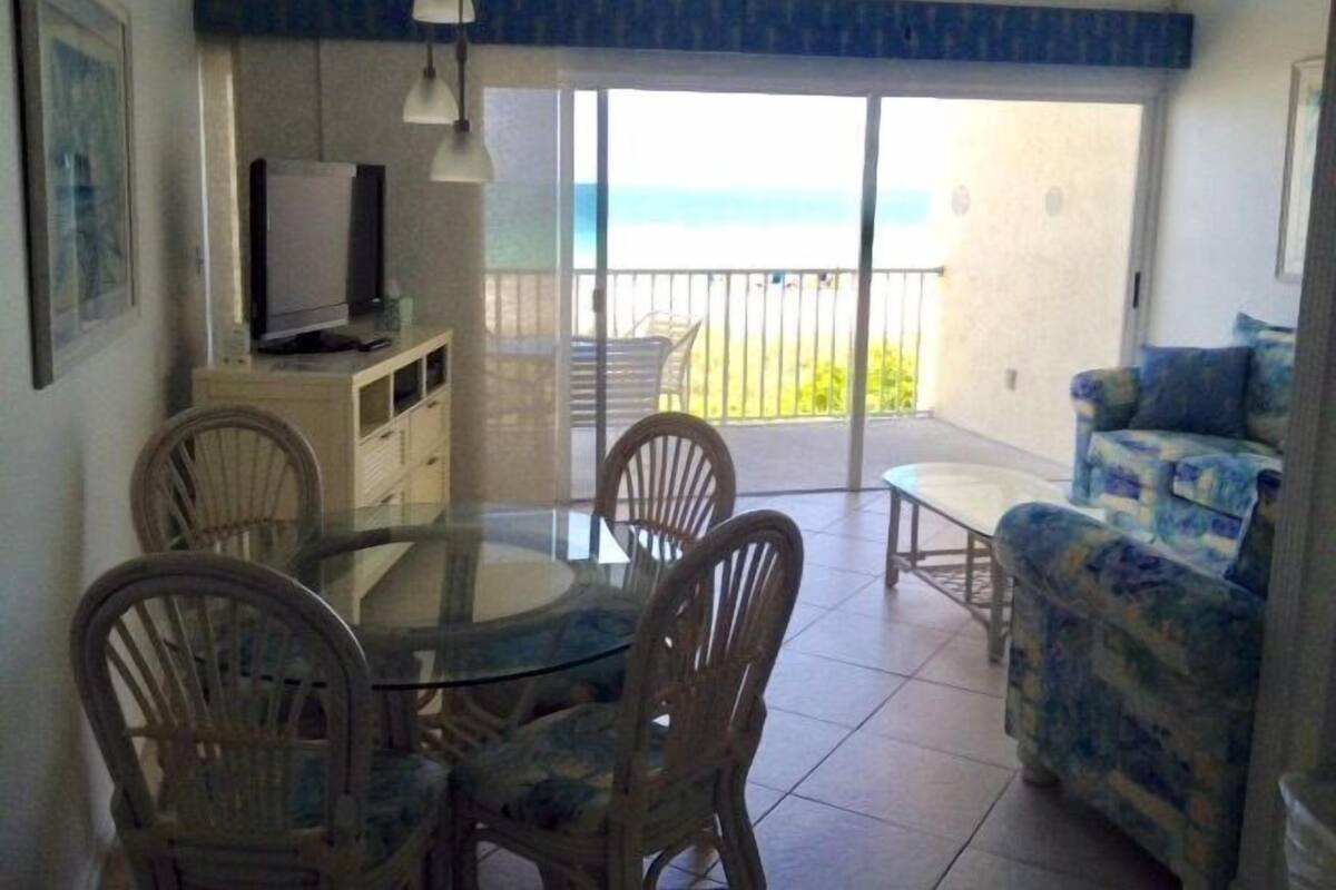 A dining area with a glass table, four chairs, a sofa, and a TV on a cabinet.