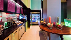 Ein moderner Frühstücksbereich mit Mikrowelle, Toaster und verschiedenen Speisen auf der Arbeitsfläche. Im Hintergrund befindet sich ein Kühlschrank und ein Tisch mit Snacks.