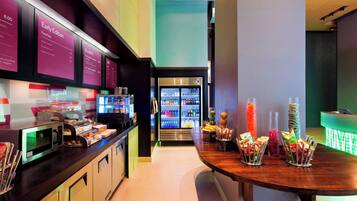 Un espace moderne dédié au petit-déjeuner, équipé d’un four à micro-ondes, d’un grille-pain et de divers aliments disposés sur le comptoir. On aperçoit un réfrigérateur au fond et une table avec des collations.
