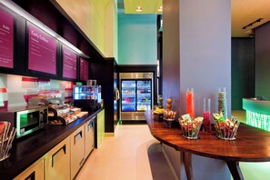 A modern breakfast area with a microwave, toaster, and various food items on the counter. There is a refrigerator in the background and a table with snacks. - Aloft by Marriott Manhattan Downtown - Financial District (New York)