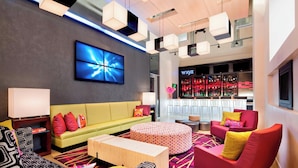 A modern hotel lobby with a yellow sofa, red armchair, and patterned ottoman. A large flat-screen TV is mounted on the wall, and a bar area with shelves of bottles is visible in the background. - Aloft by Marriott Manhattan Downtown - Financial District (New York)