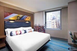Une chambre d’hôtel avec un grand lit, une vue sur la ville par la fenêtre, une tête de lit dotée d’un écran numérique et une chaise près de la fenêtre.