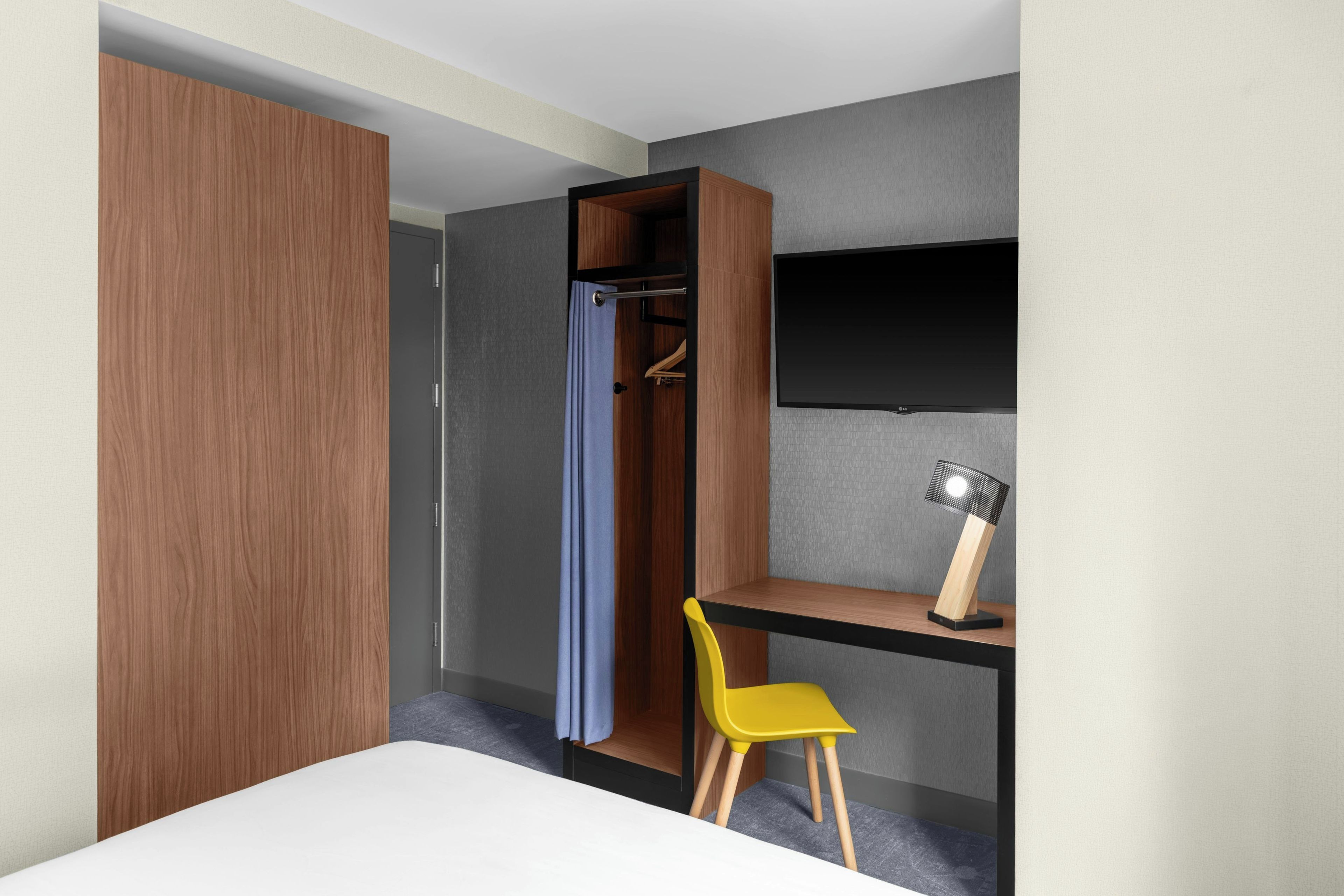 A modern hotel room with a bed, a desk, a chair, a wardrobe, a television, and a desk lamp.