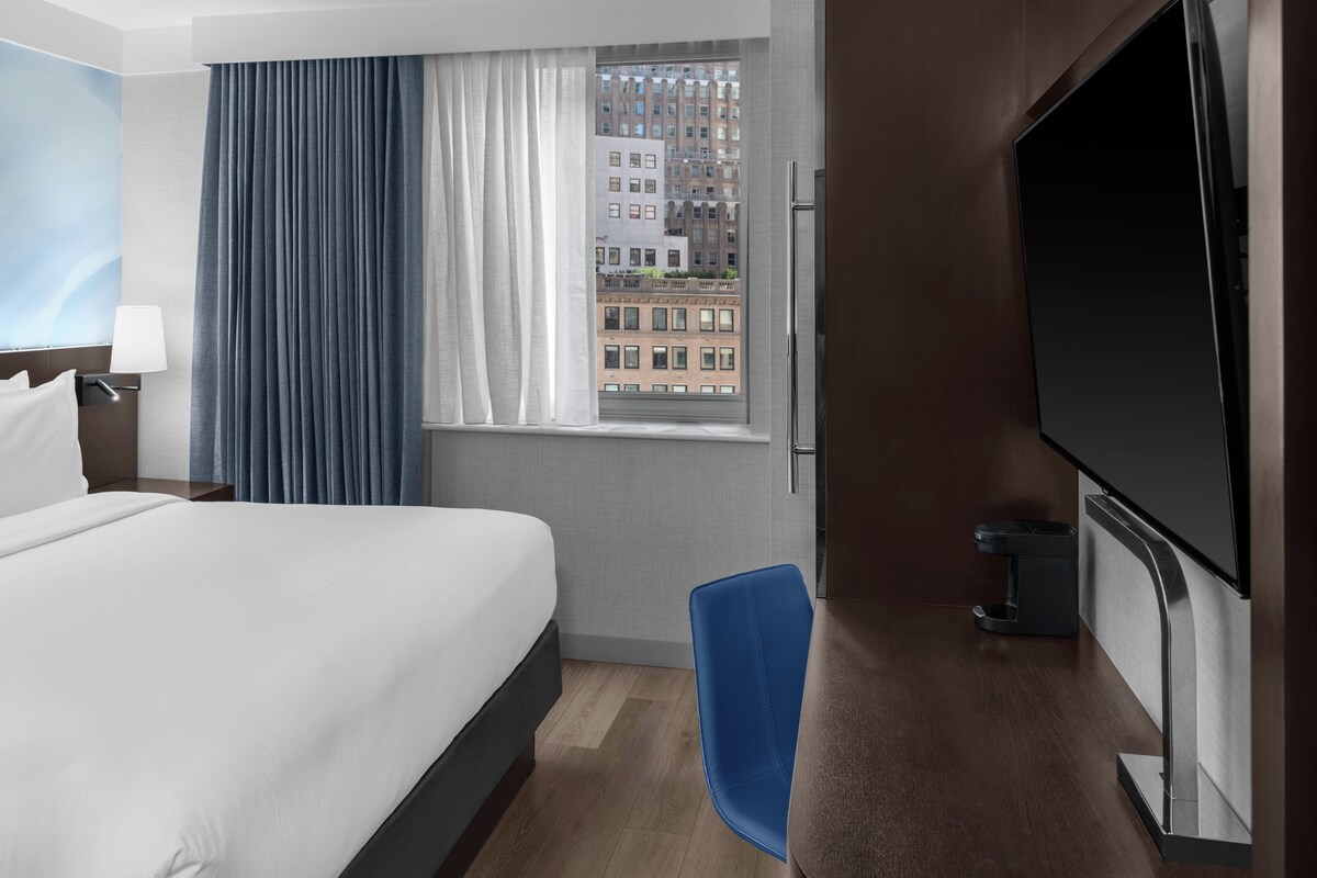 A hotel room with a bed, a desk with a TV, a chair, a window with curtains, and a view of buildings.