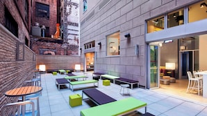 An outdoor seating area with tables and chairs, a brick wall, and a modern building entrance. - Aloft by Marriott Manhattan Downtown - Financial District (New York)