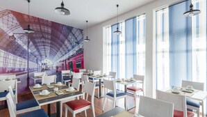 A modern dining area with white tables and chairs, a mural of a train, and large windows with blue curtains. - Brit Hotel De Paris (Saint-Jean-de-Luz)