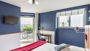 A bedroom with a bed, a desk, a chair, a TV, a window with a view of the city, and a balcony with a railing. - Brit Hotel De Paris (Saint-Jean-de-Luz)