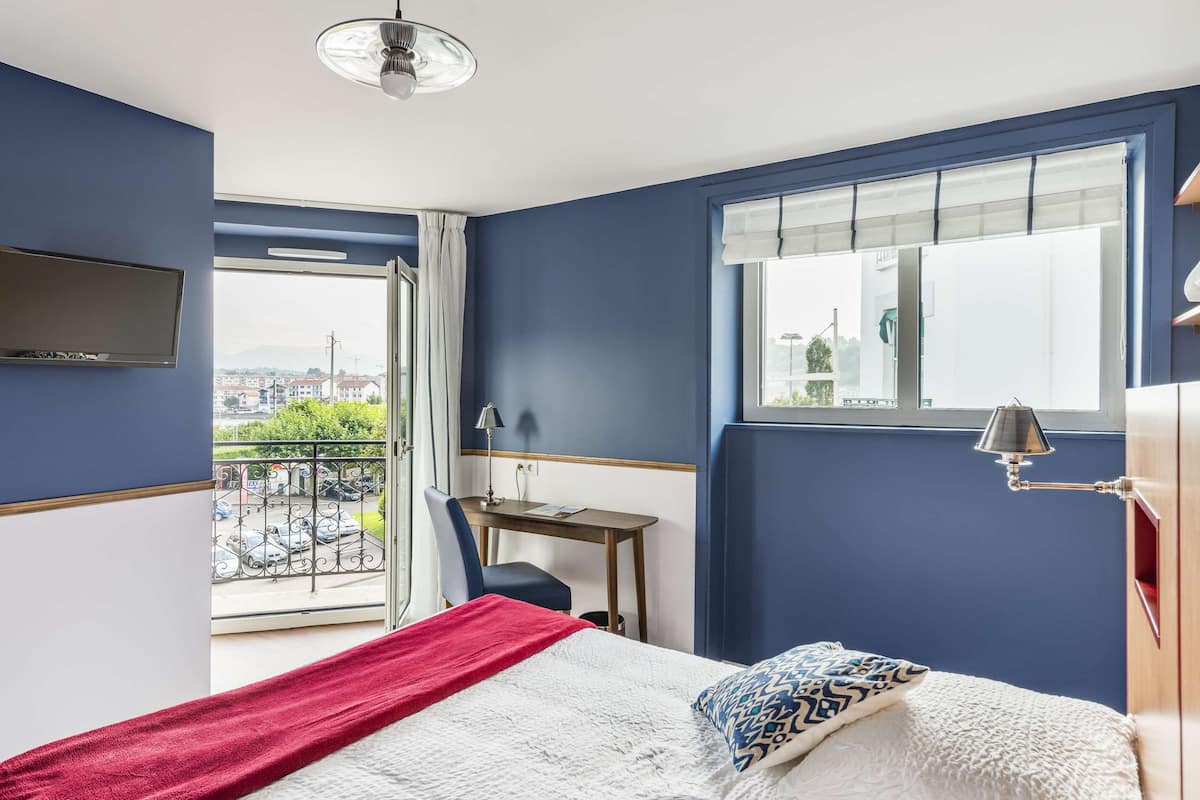 Un dormitorio con cama, escritorio, silla, televisión, ventana con vista a la ciudad y balcón con barandilla.