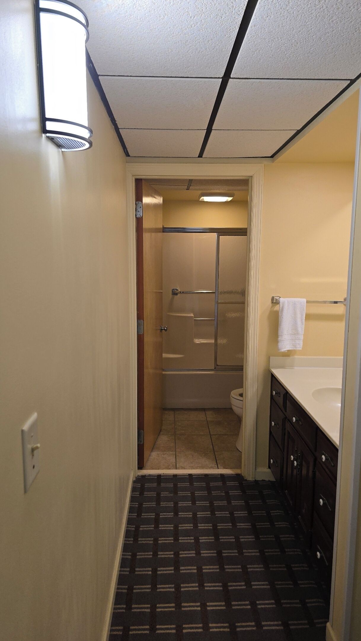 En smal gang, der fører til et badeværelse med bruser, toilet og håndvask.