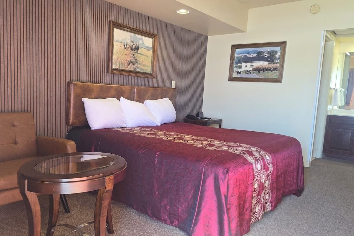 A hotel room with a large bed, a chair, a small table, and two framed pictures on the wall.