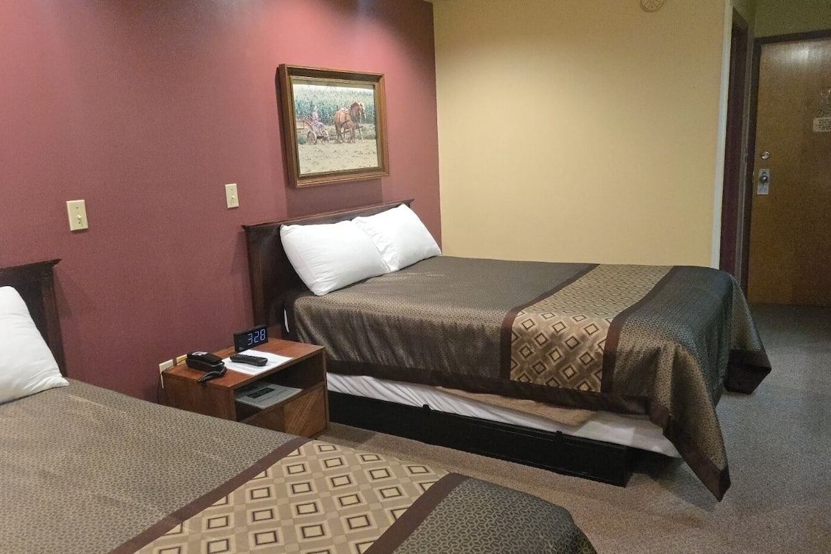 A hotel room with two beds, a nightstand with a clock, a framed picture on the wall, and a door with a sign.
