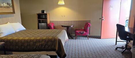 A hotel room with a bed, a desk, a chair, a small table, and a radiator.