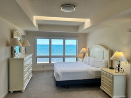 A bedroom with a large window offering a view of the sea, a bed with white linens, a dresser, and two bedside lamps.