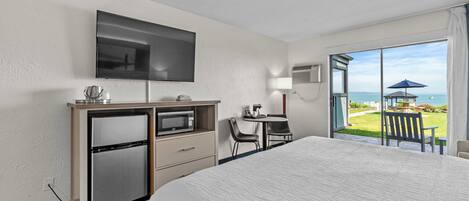 A hotel room with a bed, a desk with a chair, a television, a mini-fridge, and a view of the sea through large windows.