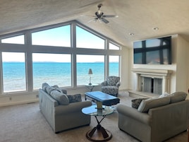 A living room with large windows overlooking the sea, a fireplace, and a ceiling fan.