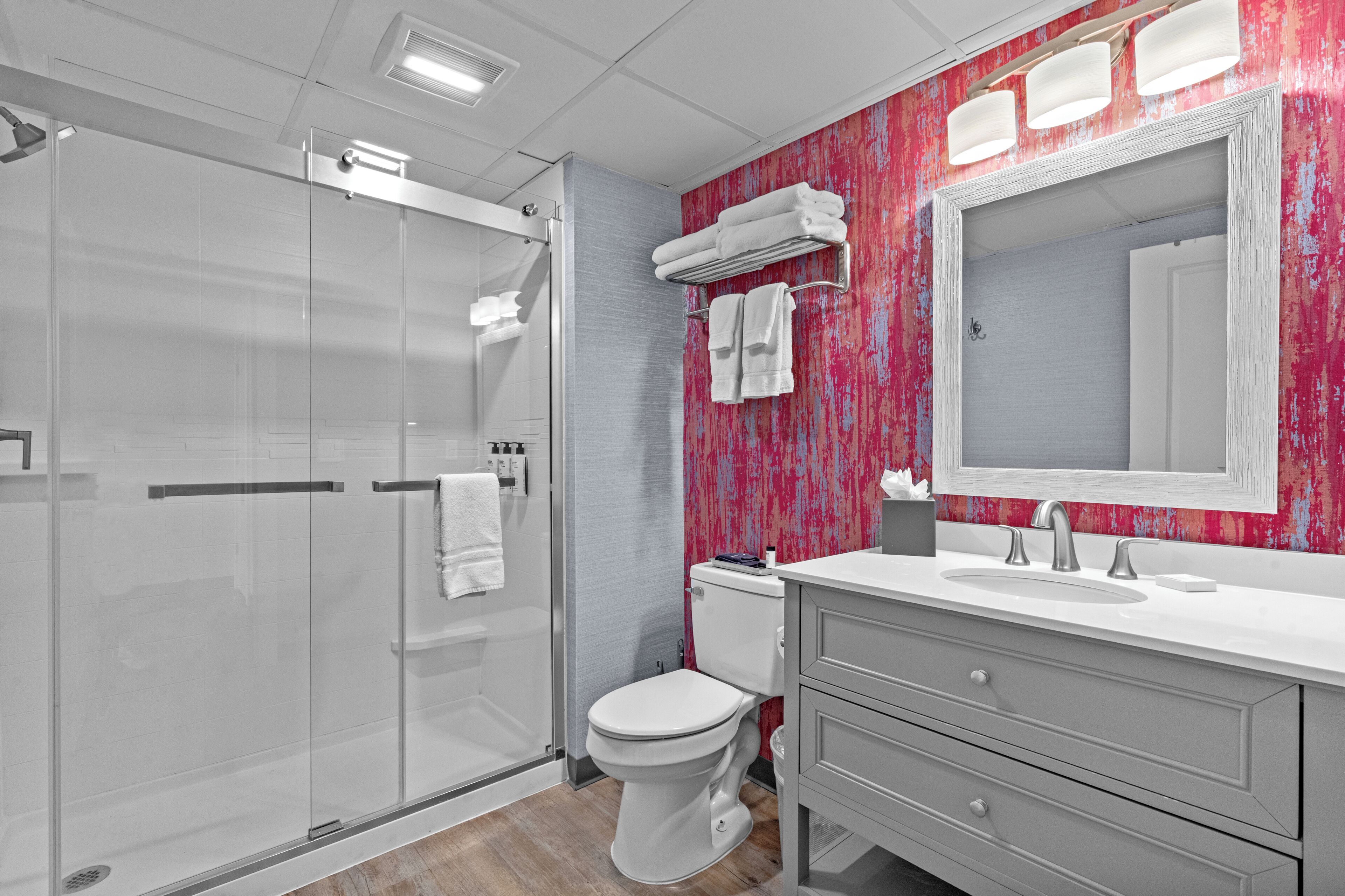 A modern bathroom with a glass shower enclosure, a white toilet, a vanity with a mirror, and a pink textured wall.