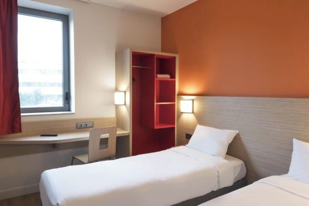 Habitación de hotel con dos camas, un escritorio con silla, un estante rojo y una ventana con cortinas.