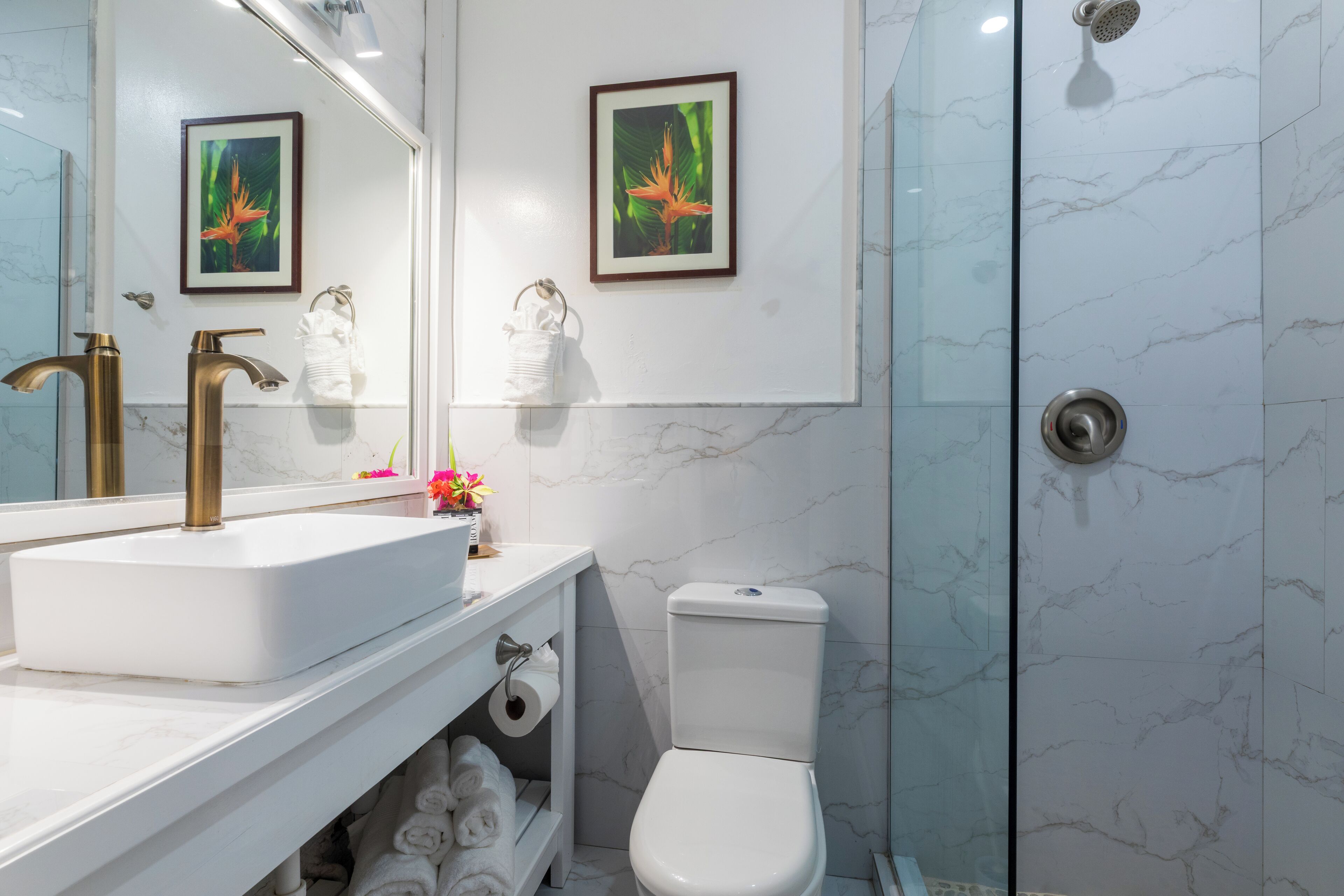 A modern bathroom with a white sink, gold faucets, a toilet, and a glass shower enclosure.