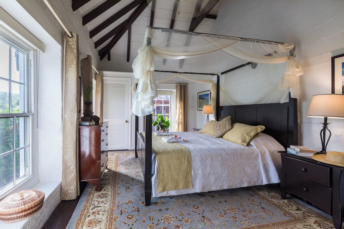 A four-poster bed with a canopy, a bedside table with a lamp, and a window with a view of greenery.