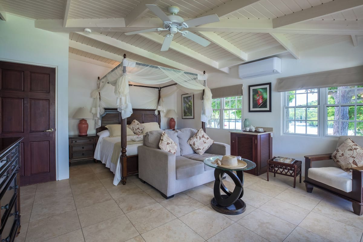 A spacious room with a four-poster bed, a sofa, and a chair. There is a ceiling fan, a window with a view of the outdoors, and a door.