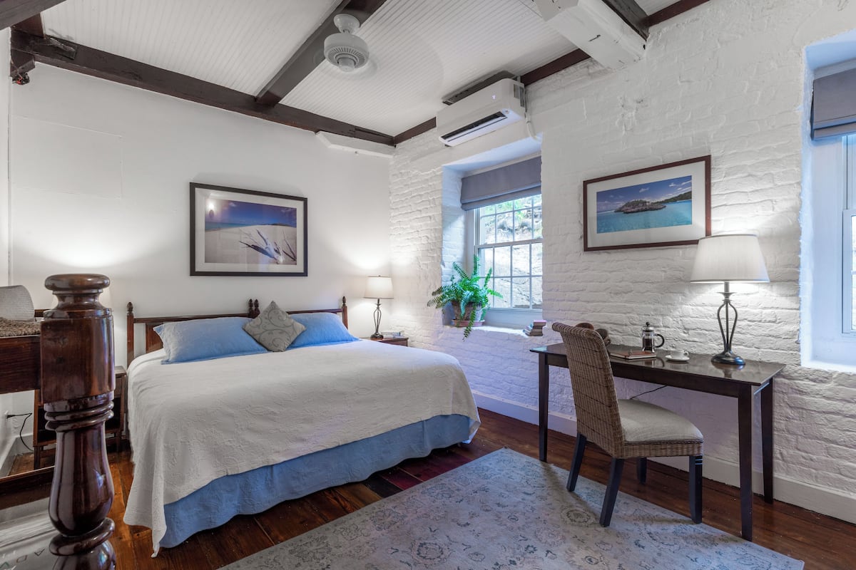 A bedroom with a large bed, a desk, and a chair. There are two windows with curtains, a potted plant, and a framed picture on the wall.