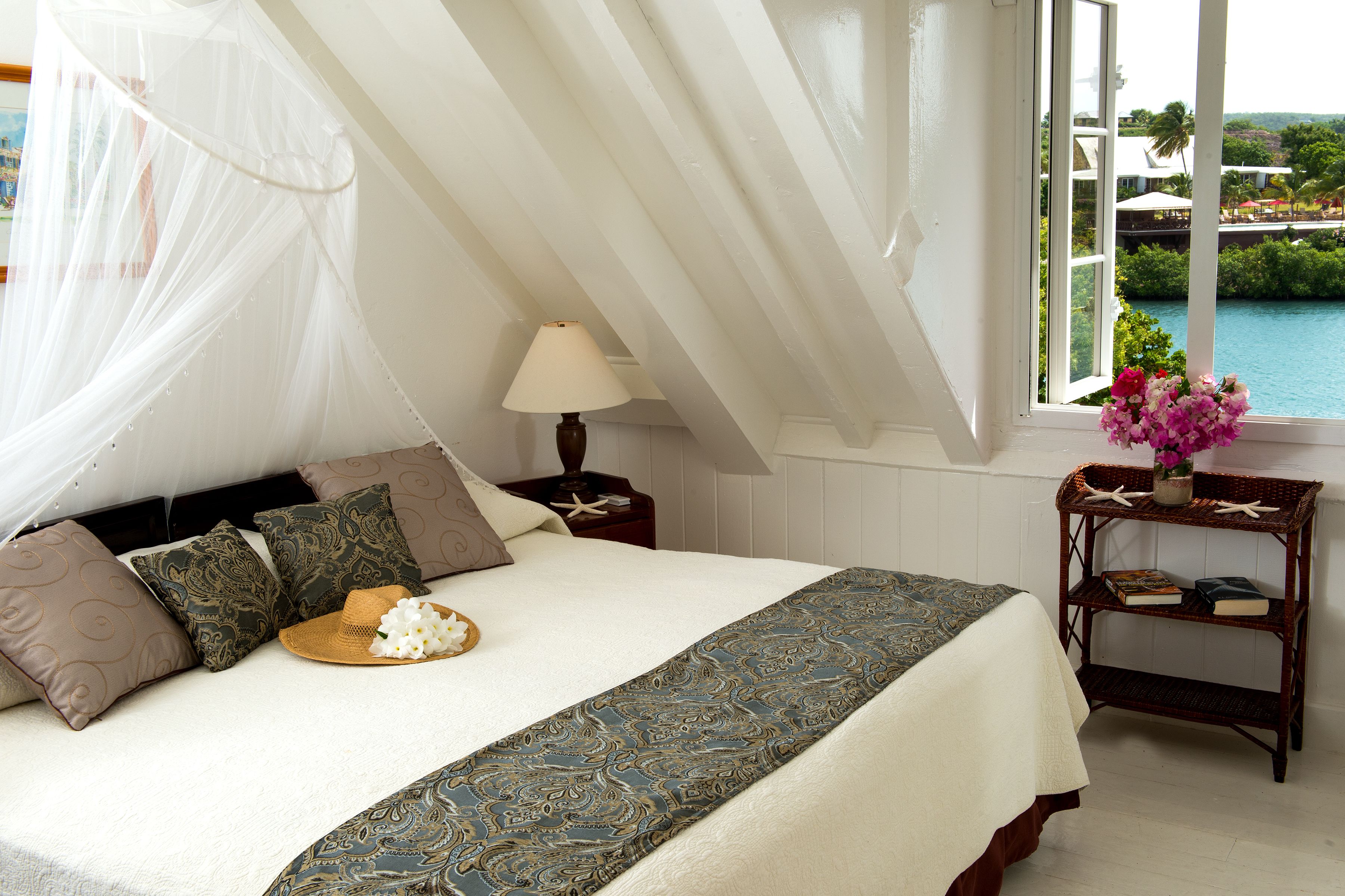 A bedroom with a bed, a nightstand, a lamp, a window with a view, and a small table with a vase of flowers.