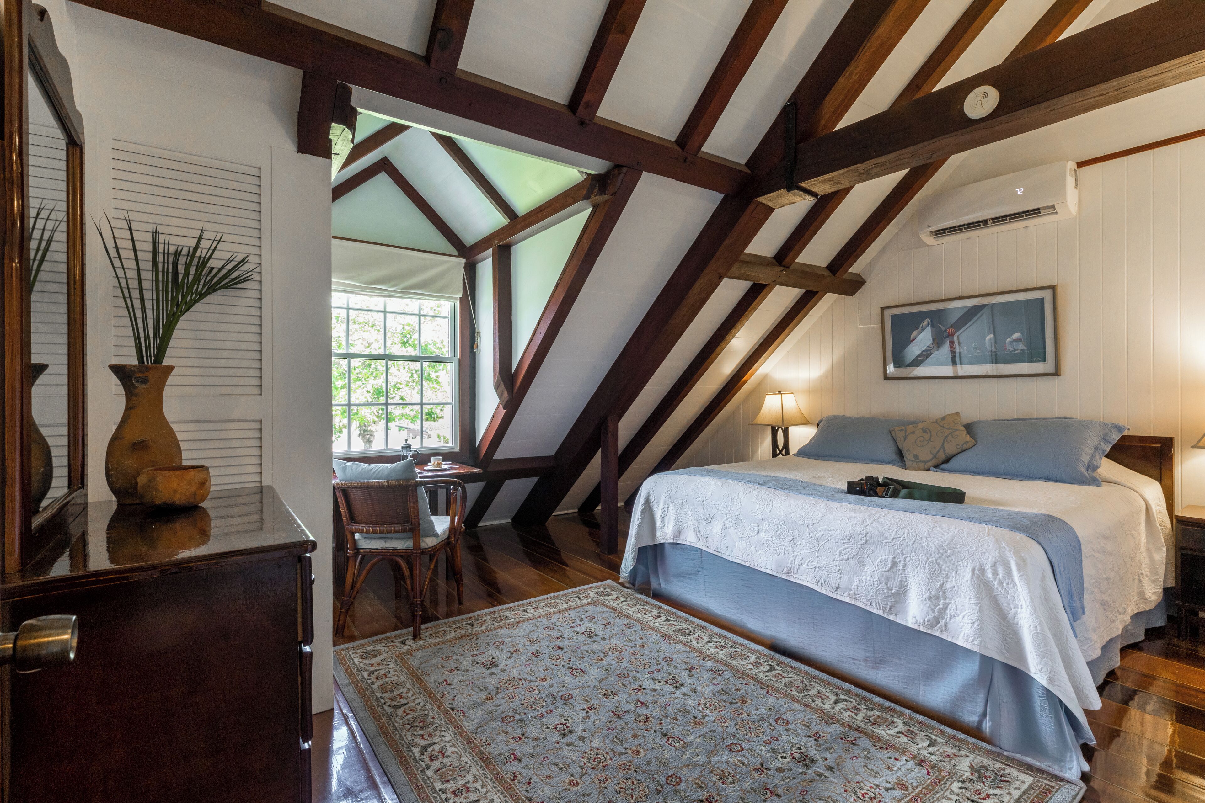 Een slaapkamer met een schuine kap, houten balken, een bed met blauw beddengoed, een nachtkastje, een lamp en een raam met uitzicht op groen.