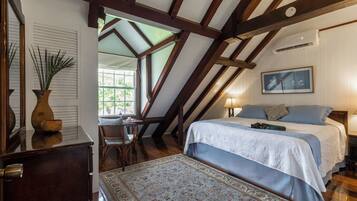 Een slaapkamer met een schuine kap, houten balken, een bed met blauw beddengoed, een nachtkastje, een lamp en een raam met uitzicht op groen.