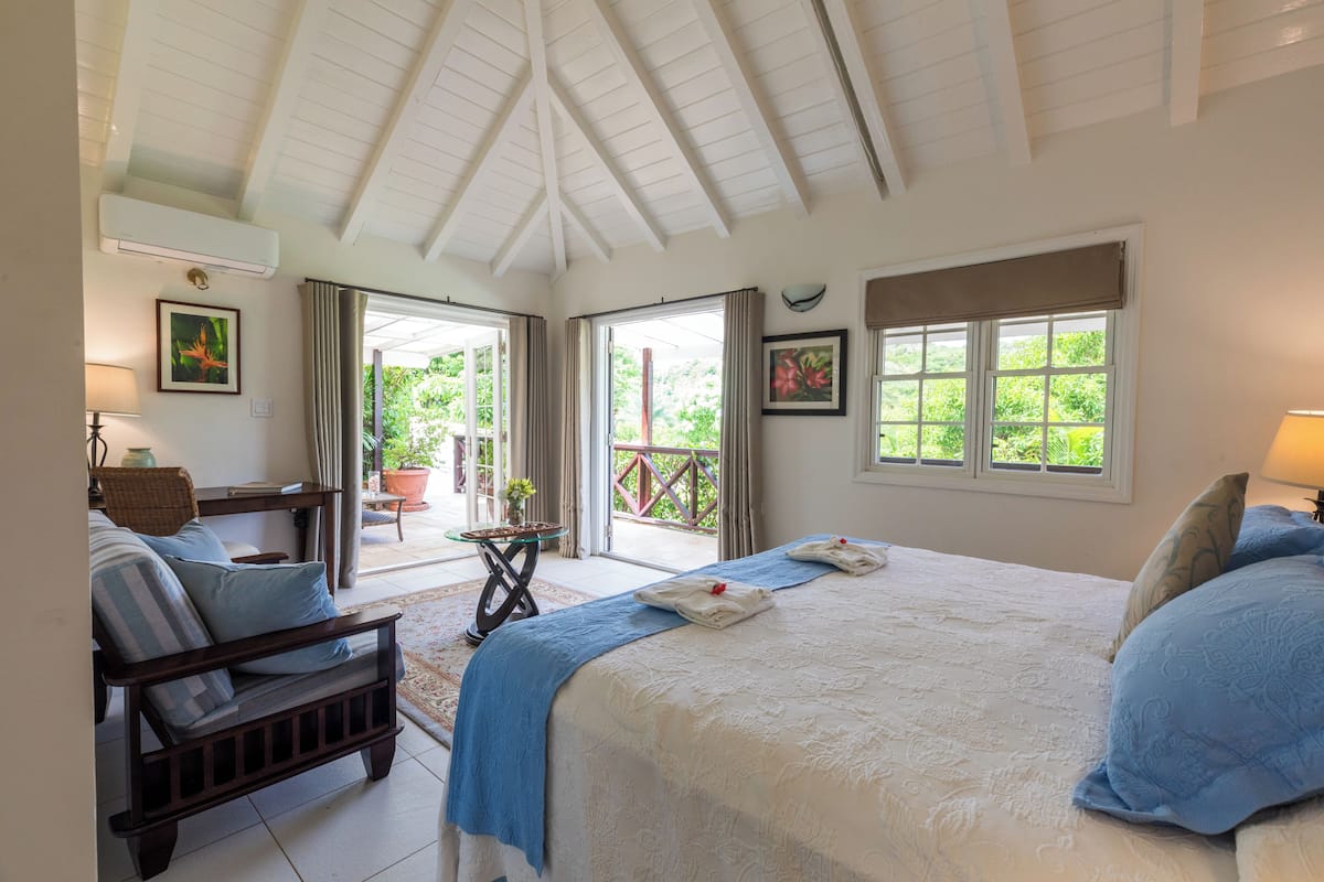 A bedroom with a large bed, a wooden bench, a table with a lamp, and a view of greenery through the windows.