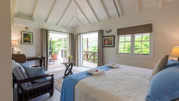 A bedroom with a large bed, a wooden bench, a table with a lamp, and a view of greenery through the windows.