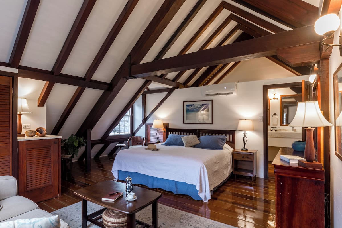 A bedroom with a sloped ceiling, wooden beams, a bed with a white bedspread, a nightstand with a lamp, and a window with a view.