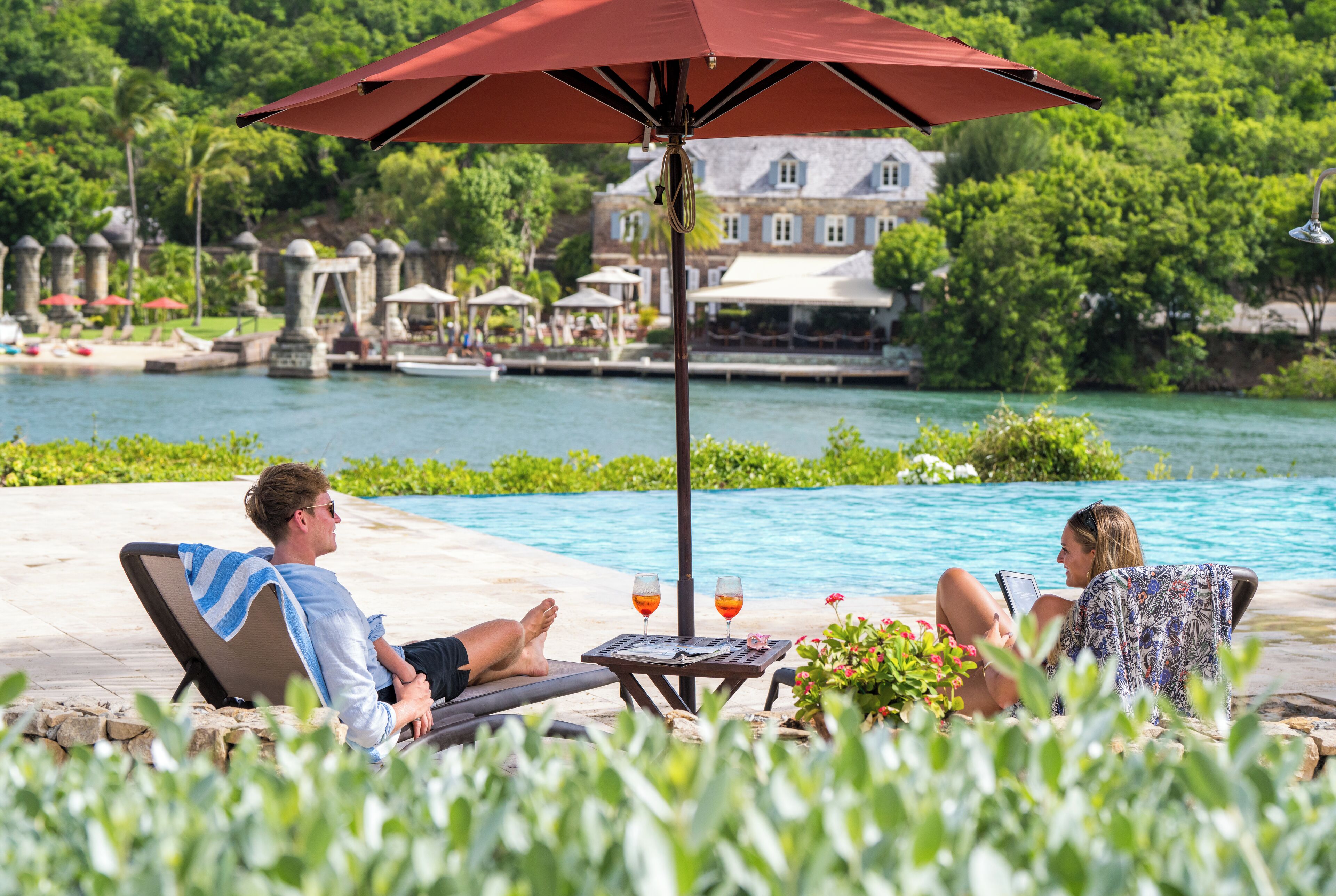 Zona de piscina con sillones de descanso, una mesa con bebidas y vista a un lago y edificios al fondo.