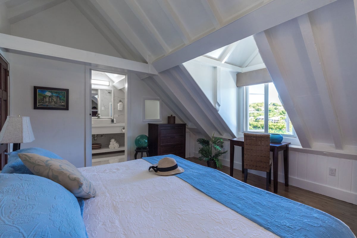 A bedroom with a large bed, a skylight, a view of greenery outside, and a small table with a chair.
