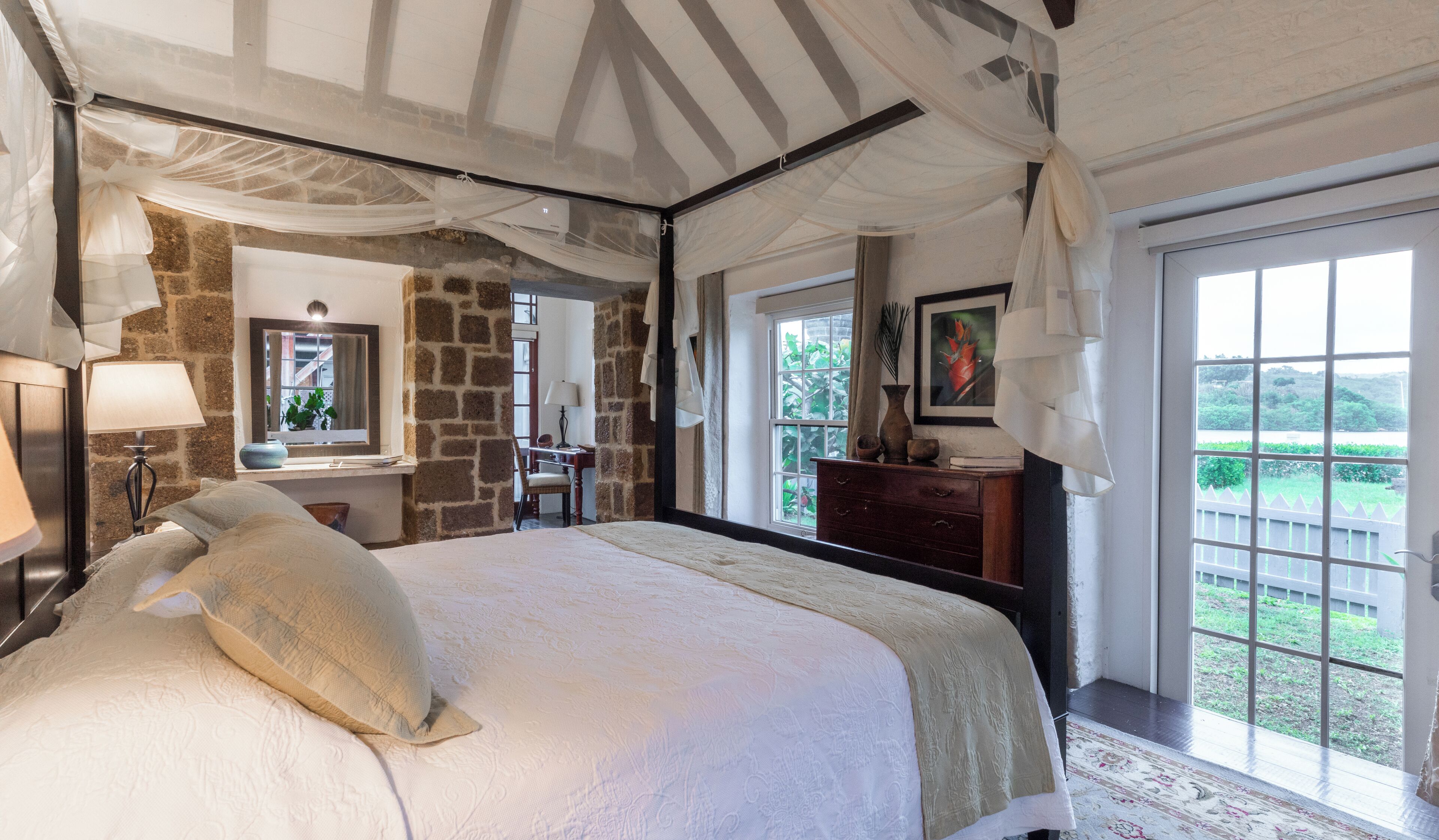 A bedroom with a large bed, a stone wall, a desk, and a view of greenery outside.