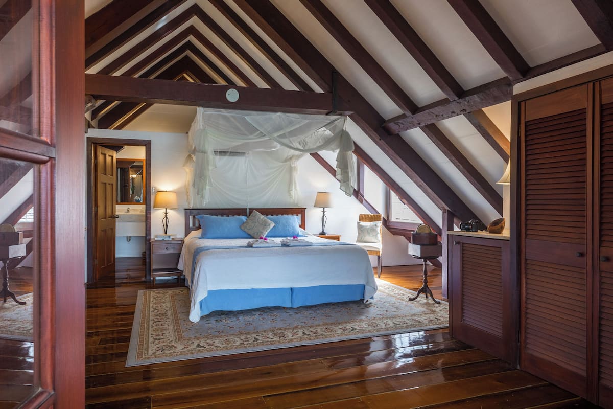 A bedroom with a wooden ceiling, a bed with blue bedding, a nightstand with a lamp, and a window with a view.