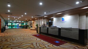 Un hall d’accueil d’hôtel avec un tapis à motifs, des comptoirs de réception portant les inscriptions « Caisse », « Enregistrement » et « Renseignements », et le logo de l’hôtel.