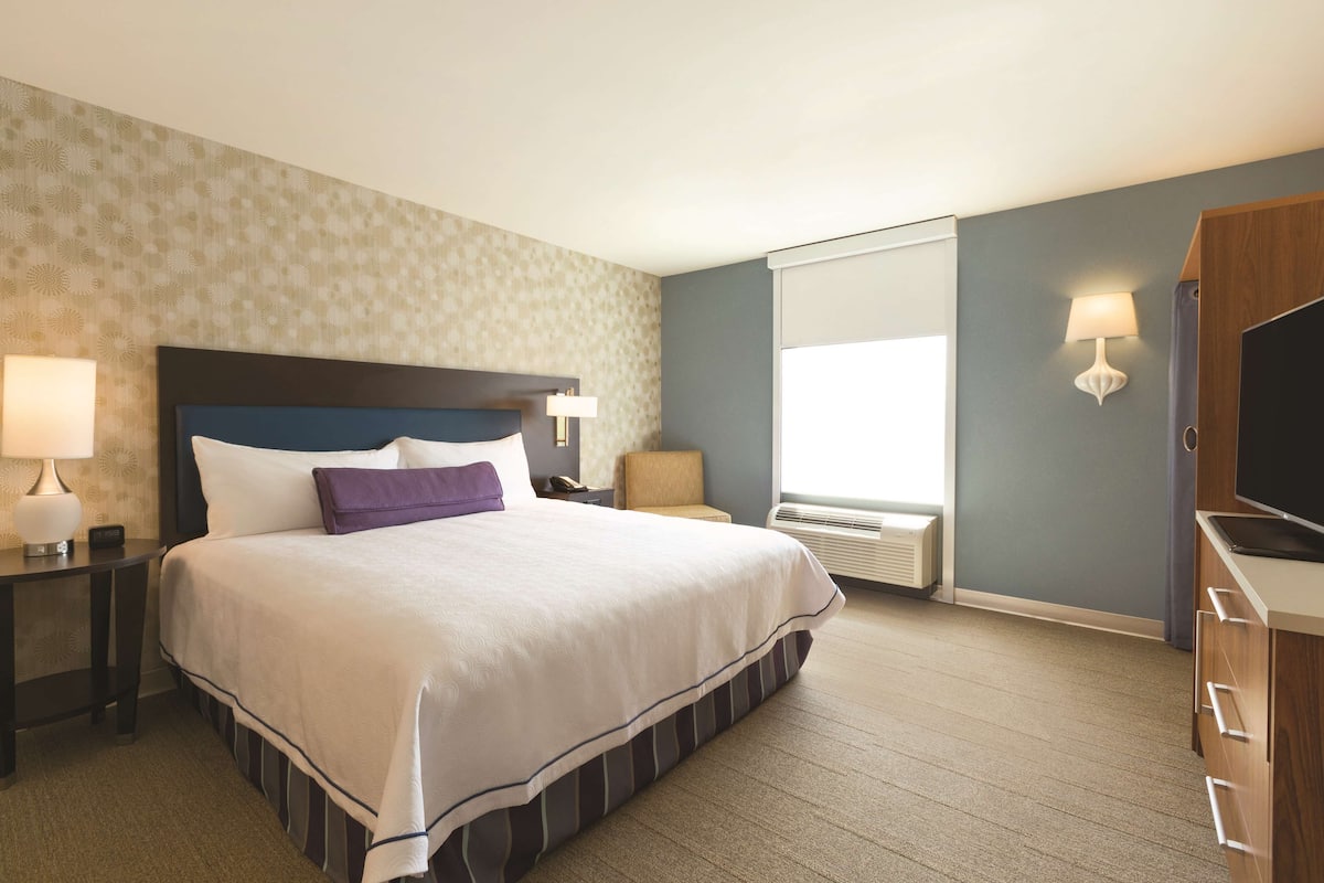 Une chambre d’hôtel avec un grand lit, une table de chevet, un bureau, une télévision et une fenêtre avec des stores.